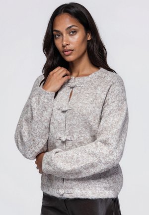 Un mannequin pose devant un fond uni, vêtu d’un cardigan gris clair texturé à fermetures brandebourg et d’un pantalon foncé.