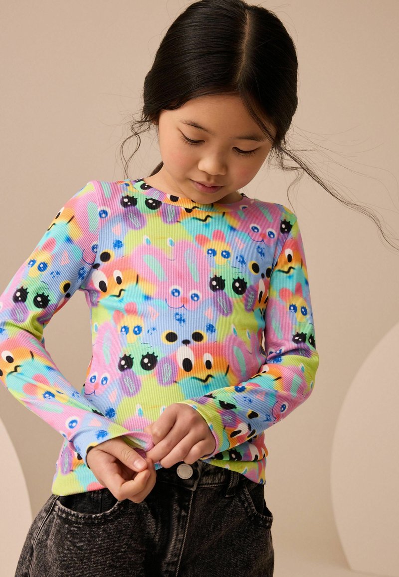 Jeune fille regardant vers le bas, portant une chemise colorée à manches longues avec des visages de dessins animés ludiques et un jean foncé sur un fond neutre.