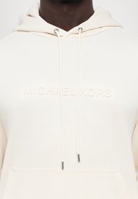 Kremet hettegenser med teksturert finish, med en lommes foran, justerbare snorer og preget 'MICHAEL KORS' logo. Sølvfarget tilbehør.