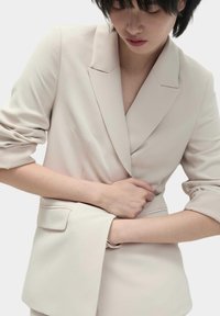 Blazer su misura beige chiaro con silhouette aderente, spalle strutturate e maniche ampie; tessuto liscio con dettagli minimi.