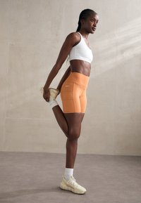 Conjunto deportivo que incluye un sujetador deportivo blanco y pantalones cortos de ciclismo de tiro alto naranjas, fabricados con un tejido suave, combinados con zapatillas amarillas claras.