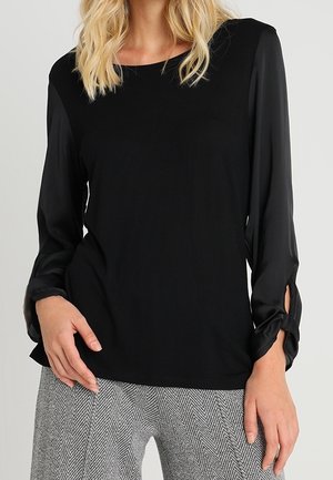 Vrouw met een zwarte blouse met lange mouwen en gestrikte manchetten, en een grijze visgraatbroek, staand met één hand nabij de taille.