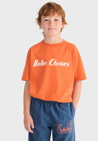 Niño pelirrojo con camiseta naranja de "Bobo Choses" y pantalones cortos azules con el texto "Pickles", de pie contra un fondo claro y liso.