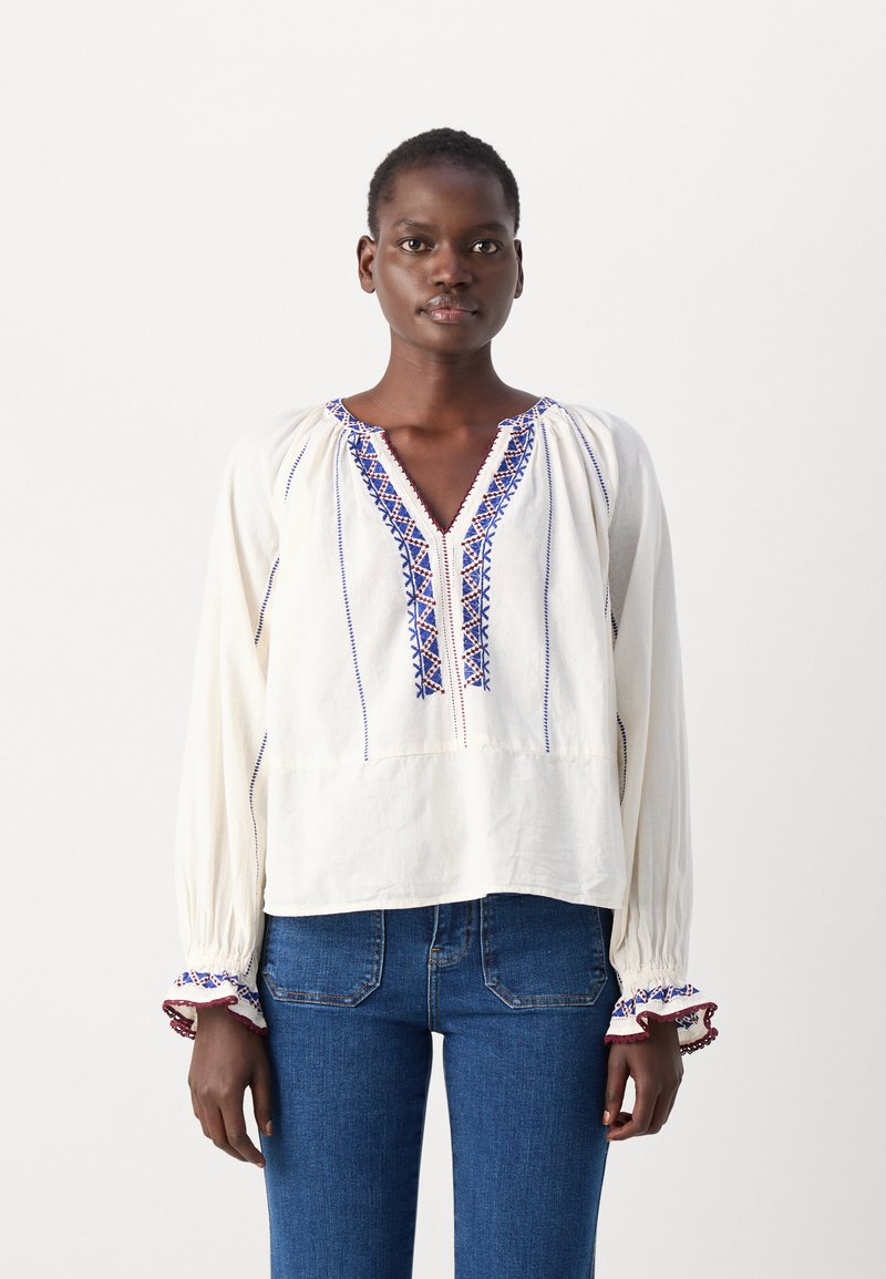 Blouse blanche brodée avec un col en V, ornée de motifs bleus et rouges le long de l'avant et des ourlets des manches. Tissu doux avec des manches longues.