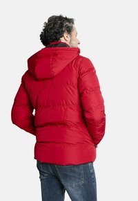 Chaqueta roja acolchada con capucha desmontable, que presenta costuras horizontales y puños elásticos, combinada con jeans azules. Tejido de poliéster texturizado.