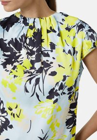 Bloes met bloemmotief in lichtblauwe stof, met levendige gele en zwarte bloemenprints, geplooide halslijn en korte mouwen.