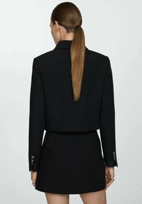 Blazer negro cropped con hombros entallados, cierre de un solo botón y mangas largas, combinado con una minifalda a juego. Textura de tela suave.