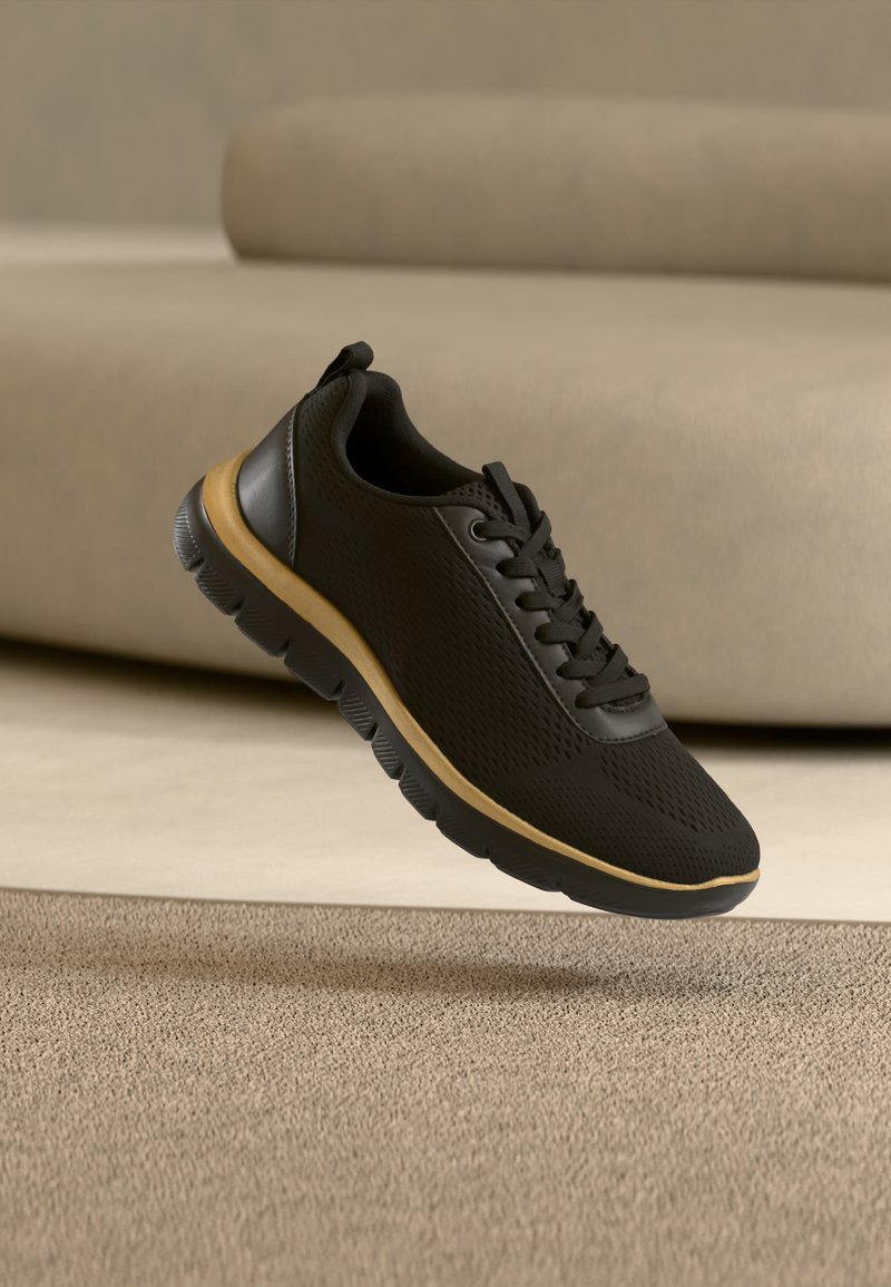 Baskets de sport noires avec semelle beige flottant au-dessus d'un tapis beige, avec un canapé beige en arrière-plan.