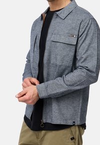 Homme portant une veste à fermeture éclair bleue sur un t-shirt noir et un pantalon beige, ajustant la manche avec les deux mains.