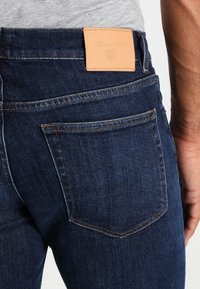 Vaqueros de mezclilla azul oscuro con una textura suave, que presentan una etiqueta de cuero marrón en la cinturilla y dos bolsillos traseros con costuras visibles.