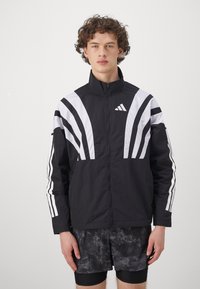 Veste de sport noire avec des rayures diagonales blanches et un logo brodé. Dispose d'un col haut et d'une fermeture éclair. Tissu texturé.
