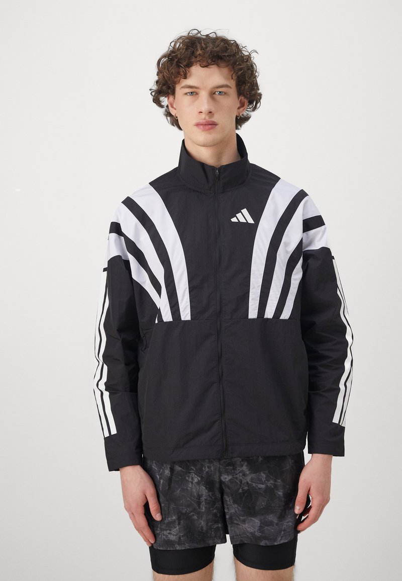 Veste de sport noire avec des rayures diagonales blanches et un logo brodé. Dispose d'un col haut et d'une fermeture éclair. Tissu texturé.