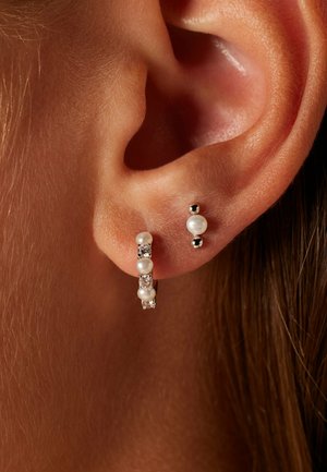 Zilveren oorring in ringvorm met parel- en kristalaccenten, naast een kleine steker met een zilveren bolletje. Close-up van oorlel.