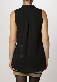 Kvinna i svart ärmlös blus med bakfåll och beige paljettkjol, sedd bakifrån mot en neutral bakgrund.