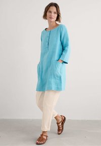 Turquoise linnen tuniek met een ronde halslijn, driekwartmouwen en twee voorzakken, gecombineerd met crèmekleurige ontspannen broek en bruine sandalen.