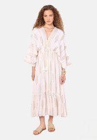 Langes, fließendes Kleid in hellem Beige mit goldenen Mustern und Quastenakzenten, mit tiefem V-Ausschnitt und weiten Ärmeln. Kombiniert mit einfachen Sandalen.