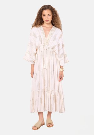 Langes, fließendes Kleid in hellem Beige mit goldenen Mustern und Quastenakzenten, mit tiefem V-Ausschnitt und weiten Ärmeln. Kombiniert mit einfachen Sandalen.