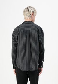 Black long-sleeve shirt featuring white vertical stripes, a button-down collar, and rounded hem. Relaxed fit with pleated details at the back.