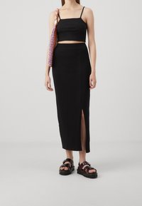 Schwarzes Crop-Top mit dünnen Trägern und einem schwarzen Midi-Rock mit einem Schlitz vorne. Kombiniert mit burgunderfarbenen Plateausandalen und einer bunten Stricktasche.