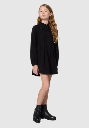Robe noire à manches longues, tissu froncé, et un motif perlé au col. Associée à des bottines noires à la cheville avec un accent doré.