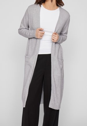 Femme portant un long cardigan gris clair avec des poches sur une chemise blanche et un pantalon large noir, les mains tenant le cardigan ouvert.