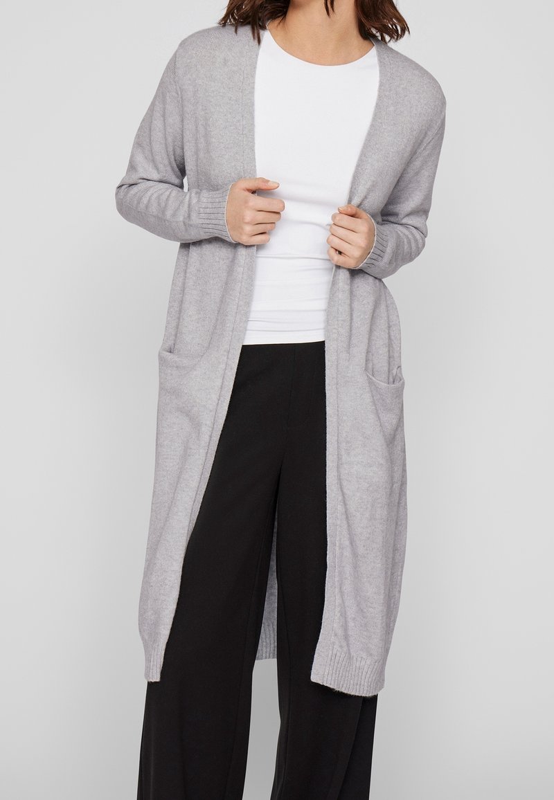 Femme portant un long cardigan gris clair avec des poches sur une chemise blanche et un pantalon large noir, les mains tenant le cardigan ouvert.
