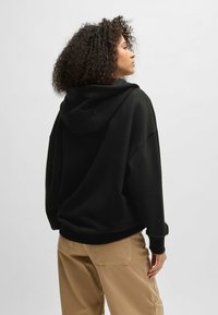 Felpa nera oversize con cappuccio regolabile, texture morbida e polsini a costine, abbinata a pantaloni cargo beige. Vista posteriore mostrata.