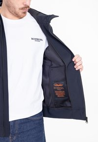 Marine bomberjack met een soepele textuur, binnenzak en een oranje label met merknaam en onderhoudsinstructies; gedragen over een wit T-shirt.