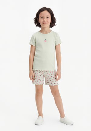 Ung pige med kort mørkt hår iført lysegrøn kortærmet top med jordbær-patch og matchende shorts med jordbærprint og hvide sko.