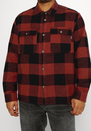 Homme portant une chemise à carreaux rouge et noire boutonnée avec deux poches poitrine et un jean gris foncé.