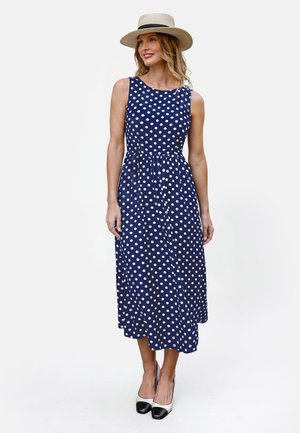 Donna che indossa un vestito midi senza maniche blu navy con pois bianchi, un cappello di paglia e tacchi bicolore neri e bianchi, in piedi su sfondo bianco.