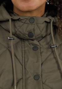 Kaki takkikera takki korkealla kauluksella, jossa on mustia neppareita, nyörejä ja pehmeä sisävuori. Huomattava timanttikuvio kankaassa.