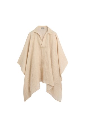 Frau geht drinnen und trägt einen lockeren beigen Poncho, dunkelbraune Hosen, schwarze Sandalen und hat ihre Haare zu einem niedrigen Dutt zurückgebunden.