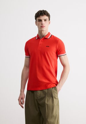 Homme portant un polo rouge avec un col et des bords de manches rayés, associé à un pantalon ample vert olive, debout devant un fond blanc.