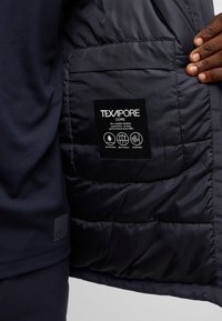 L'intérieur de la doudoune noire présente une étiquette TEXAPORE, indiquant des matériaux imperméables, respirants et coupe-vent. Texture lisse, design matelassé.