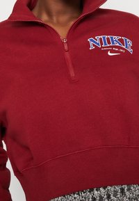 Czerwony sweatshirt z krótkim zamkiem i ściągaczem, zdobiony niebieskim i białym logo "NIKE", wykonany z miękkiego materiału. Zamek na kołnierzu i minimalne akcenty.