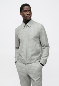 Γκρι σακάκι με λεπτές ρίγες, γιακά, φερμουάρ και δύο πλαϊνές τσέπες. Το ύφασμα έχει λεπτή υφή και φοριέται πάνω από μαύρο τοπ.