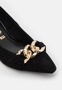 Chaussure plate en daim noir à bout pointu avec une large chaîne dorée brillante en décoration sur l\'avant.