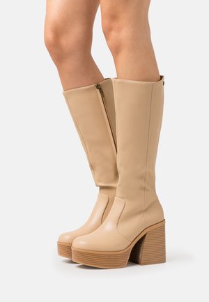 Bottes beige montantes à plateforme avec talons en bois épais, fermetures éclair latérales et finition en cuir lisse portées sur des jambes sur un fond uni.