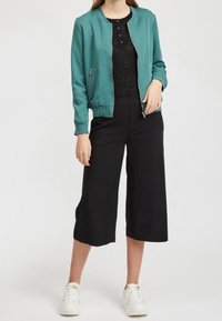 Femme portant une veste zippée bleu sarcelle, pantalon large noir court, top noir et baskets blanches, debout devant un fond blanc.