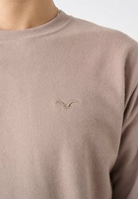 Light beige sweater with a round neck and long sleeves. Features a subtle embroidered logo on the chest. Soft, textured fabric.