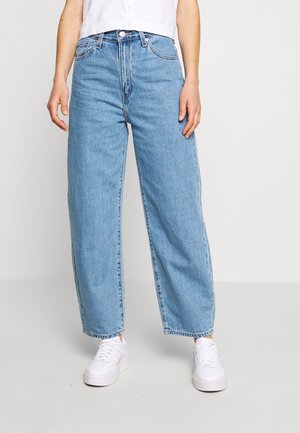 Person iført lyseblå, højtaljede, lige ben jeans og hvide sneakers, stående mod en ensfarvet, lys baggrund.