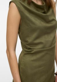 Robe sans manches verte olive avec un col drapé, présentée sur une personne, de l'épaule jusqu'à la mi-cuisse, sur un fond uni.