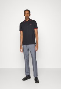 Polo shirt nera con colletto e dettagli rossi, logo sul petto, abbinata a pantaloni grigi e scarpe nere. Design minimalista, texture liscia.