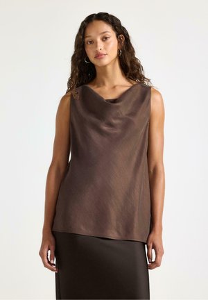 Kvinde med krøllet hår iført ærmeløs brun draperet top og matchende nederdel mod ensfarvet lys baggrund.