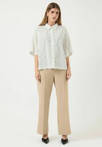 Blusa bianca a occhiello con colletto e maniche corte, abbinata a pantaloni beige a gamba larga e scarpe nere a punta.