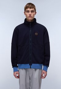 Marineblå zip-up jakke med høy krage og blå detaljer. Har frontglidelås, sidelommer, og et rundt logo-patch på brystet.