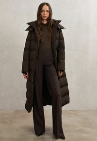 Lungo piumino marrone scuro con colletto alto e chiusure a scatto, indossato sopra pantaloni marrone scuro aderenti a vita alta. Tessuto liscio, design minimalista.