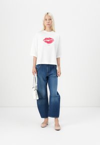 Weißes Kurzarmhemd mit rotem Lippen-Graphic, kombiniert mit weit geschnittenen blauen Jeans. Das Model hält eine kleine weiße Handtasche und trägt helle Ballerinas.