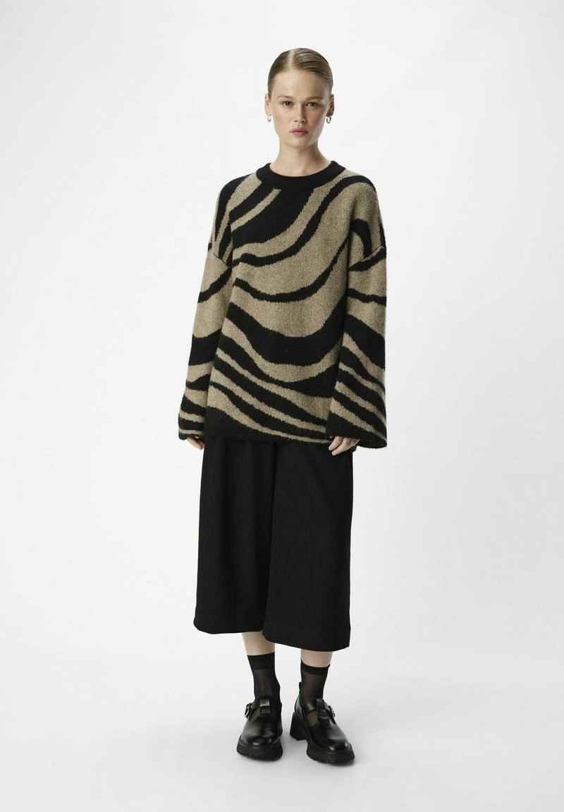 Beige Strickpullover mit schwarzen wellenförmigen Streifen, Rundhalsausschnitt und überschnittenen Schultern, kombiniert mit lockeren schwarzen Culottes und klobigen Schuhen.